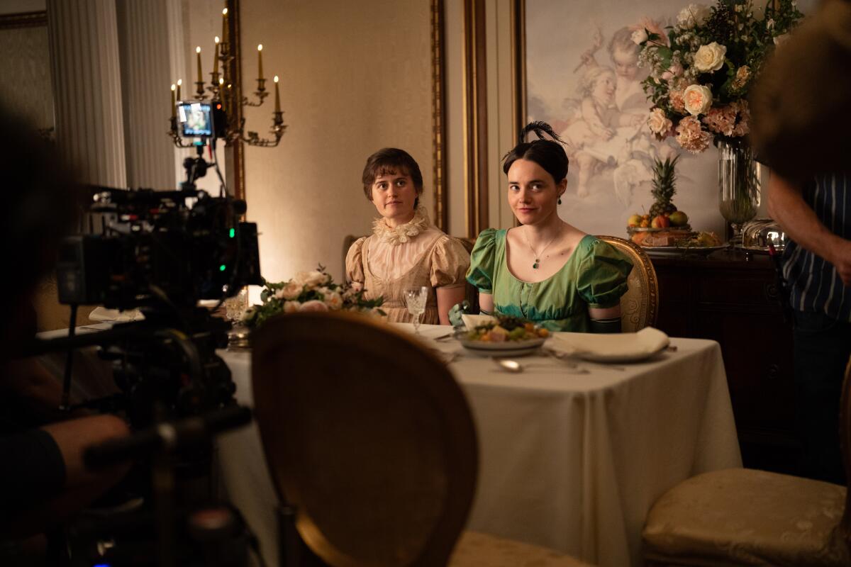 Two women in period dress sit at a table as a camera is seen before them.