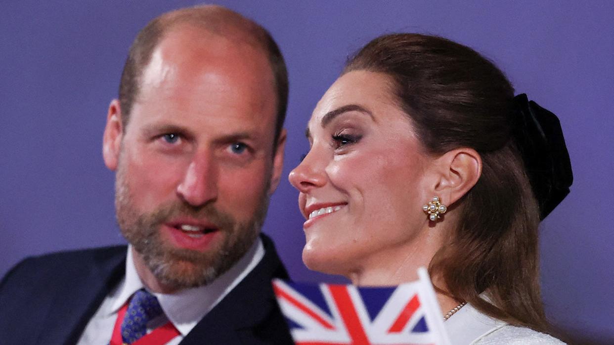 Prince William speaking to Catherine, Princess of Wales, with a Union Jack flag in front of her