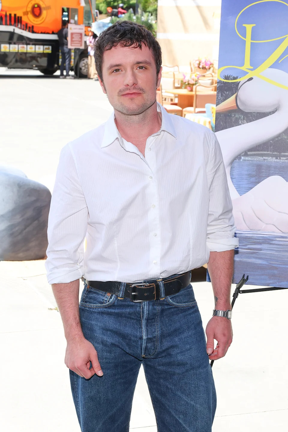 Person in a casual white shirt and jeans at an outdoor event, standing in front of a poster