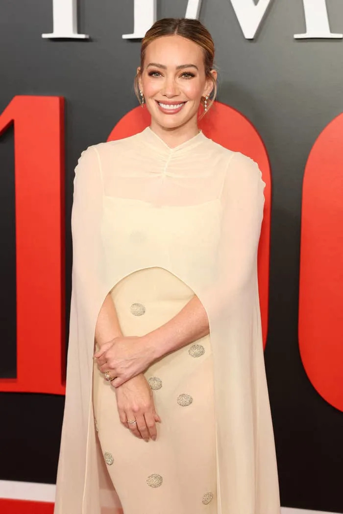 A woman on a red carpet wearing an elegant, sheer cape dress with embellishments, smiling for the camera