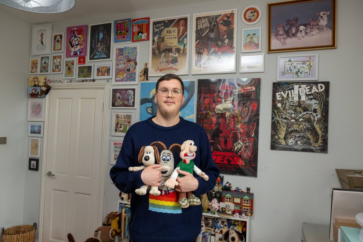 An individual stands in an interior space adorned with an array of framed images on the wall. The person holds two plush toys in their hands, dressed in a long-sleeved sweater.