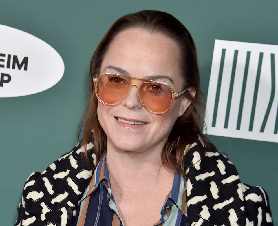 Taryn Manning wearing sunglasses and a patterned coat over a striped shirt, standing in front of a step-and-repeat backdrop