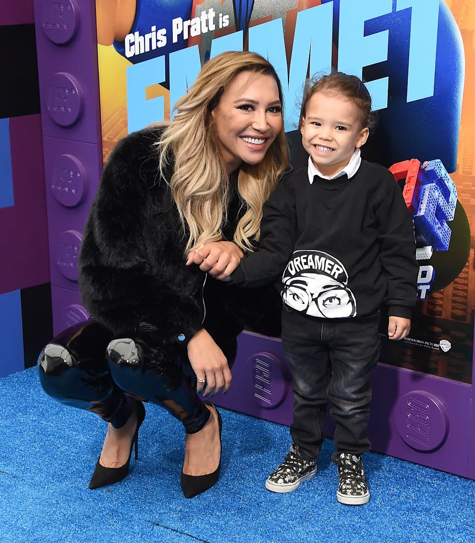 Person smiling and crouching next to a child at a movie event. The adult is wearing a fur-style jacket and shiny boots, while the child wears a graphic sweater