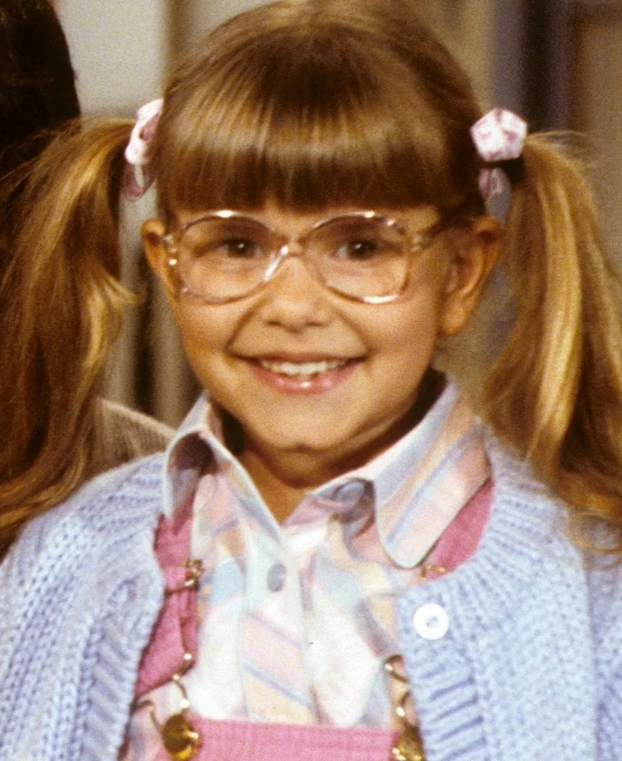 Judith Barsi in a striped shirt, overalls, and a knit sweater with glasses and her hair in two pigtails