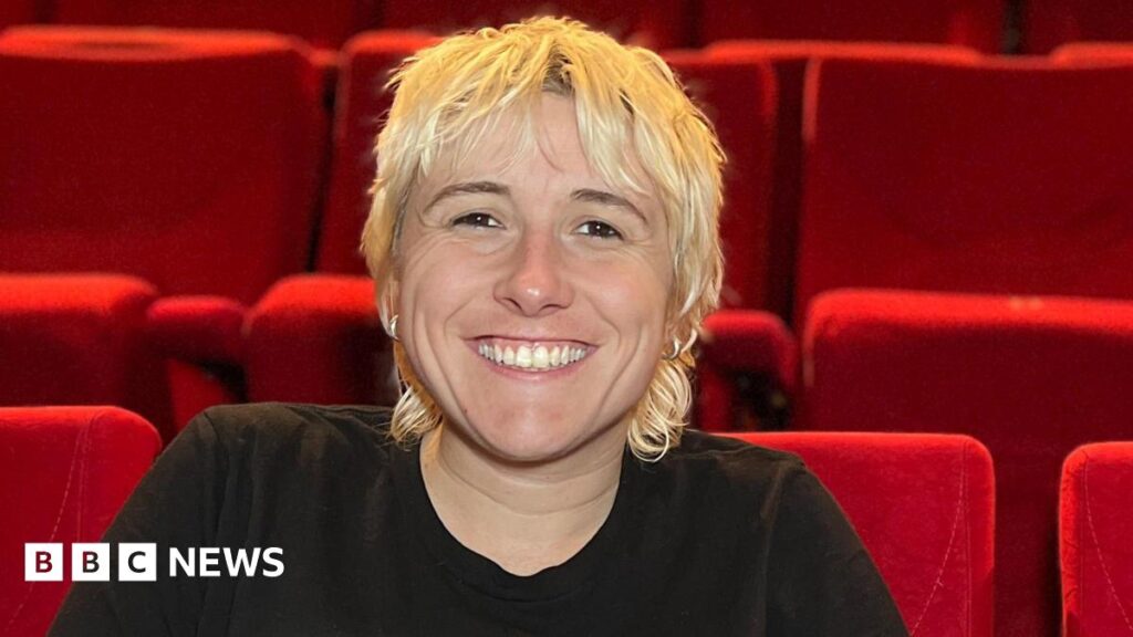 Maisie has short, shaggy blond hair and sits smiling in a theatre auditorium, leaning forward with hands clasped. She is wearing a black T‑shirt and sits among rows of bright red seats that fill the background.