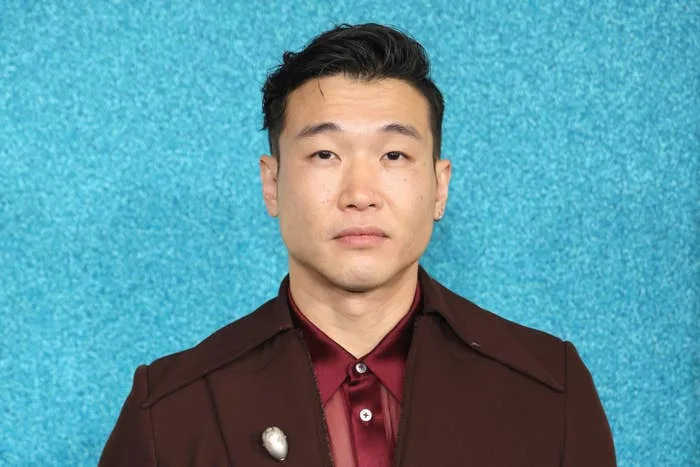 Joel Kim Booster in a formal red shirt and a brown blazer with a brooch, posing against a textured background
