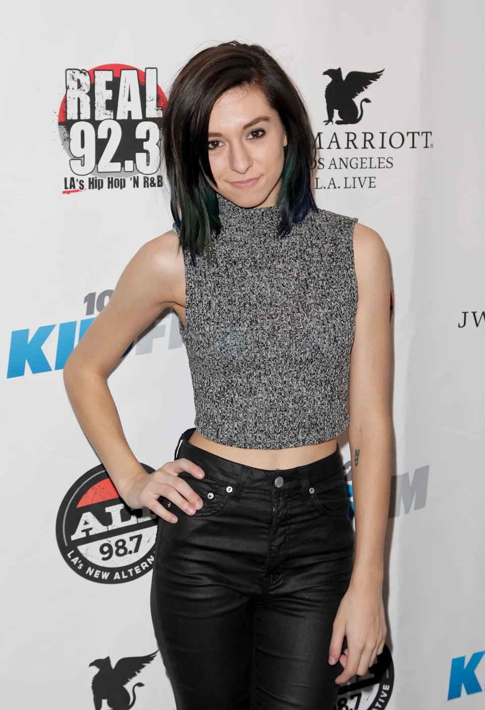 Christina Grimmie posing in a sleeveless glittery top and high-waisted pants at an event backdrop with radio station logos