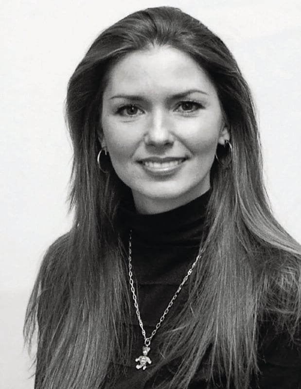 A woman with long straight hair, wearing hoop earrings, a dark turtleneck, and a necklace with a pendant, smiles at the camera against a plain background. The image is in black and white.