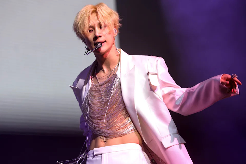 Taemin in an open suit jacket with a chain-like top, performing energetically on stage with a microphone headset