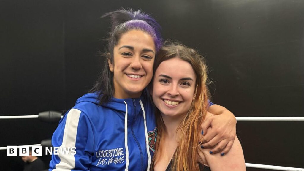 Two woman - Bayley and Natasha Swann - smile for the camera while standing in a wrestling ring. Bayley is wearing a blue tracksuit top saying Lodestone Las Vegas and has dark hair, partly dyed purple. Natasha is shorter, with red hair going past her shoulders.
