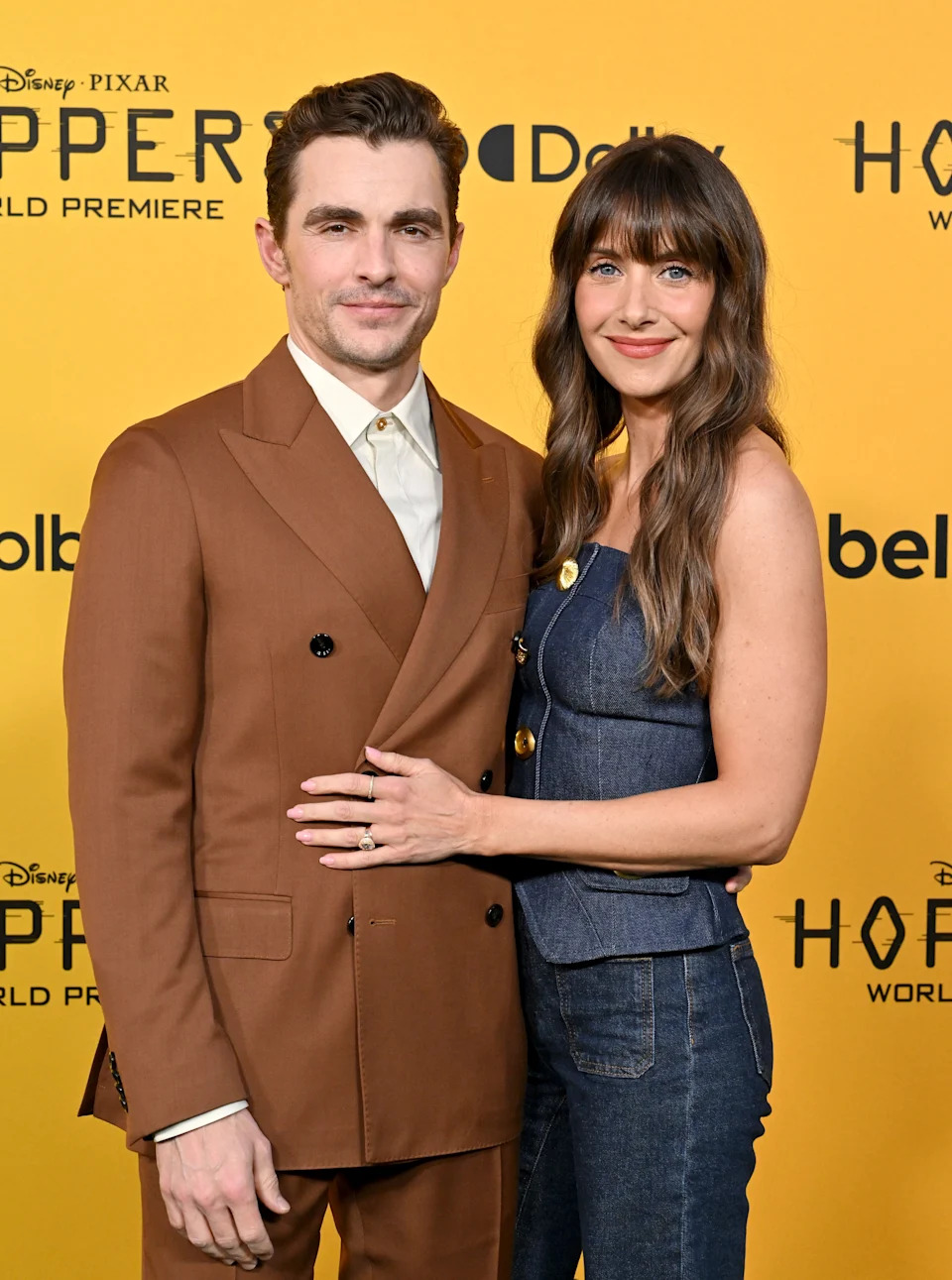 A man in a brown suit and a woman in a denim dress pose on the red carpet at a film premiere