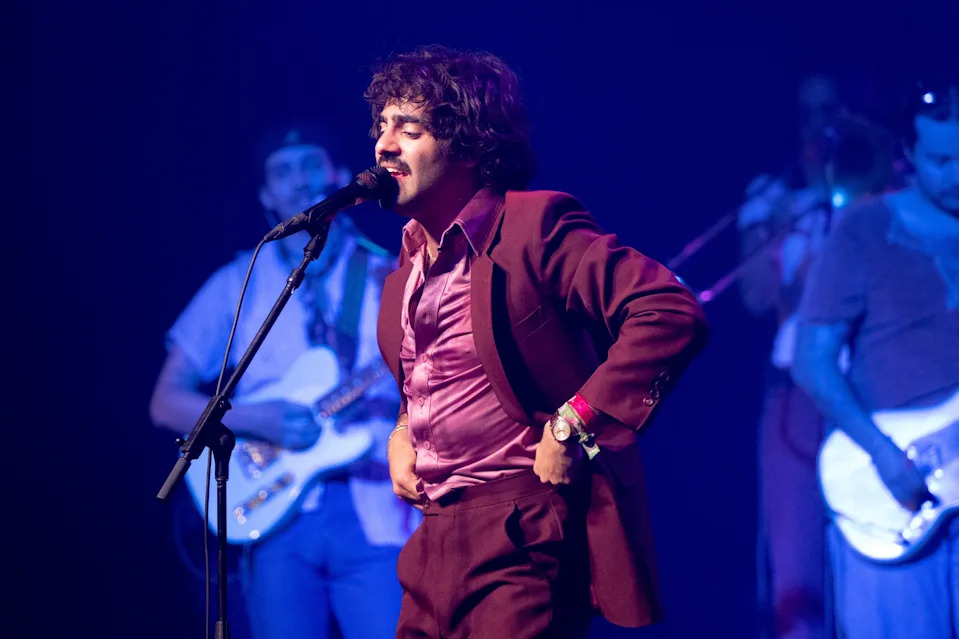 54 Ultra on stage in a vintage-inspired suit singing into a microphone, with a band playing instruments in the background