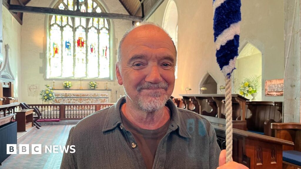 Guy Pratt stood in a cotton overshirt holding a rope connected to one of the church bells. He has a large stain glass window and the altar behind him