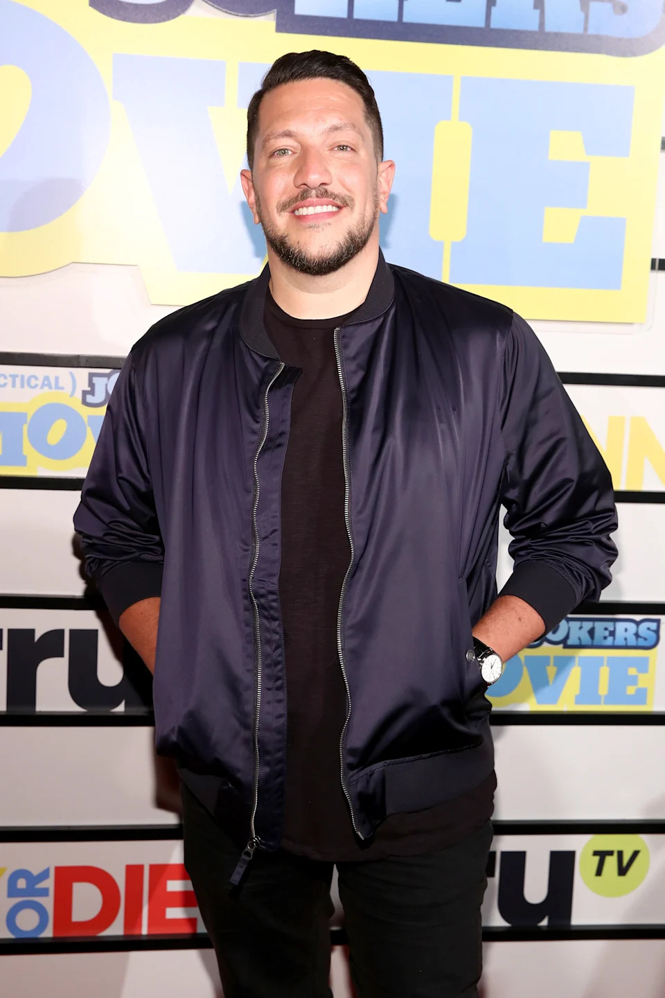 Person smiling at a movie premiere, wearing a dark bomber jacket over a t-shirt, hands in pockets, in front of a step and repeat banner