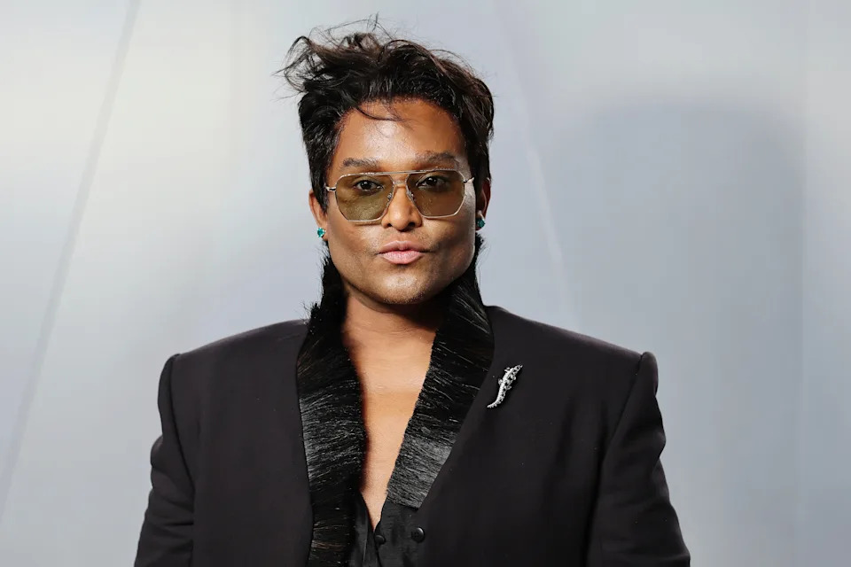 Celebrity poses confidently on a red carpet, wearing a sleek black suit with a textured collar and aviator sunglasses