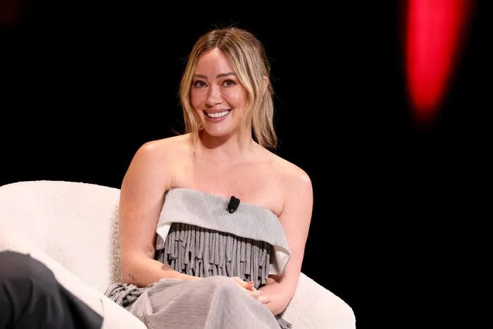 A woman in a strapless pleated dress sits on a white chair, smiling and speaking into a lapel microphone