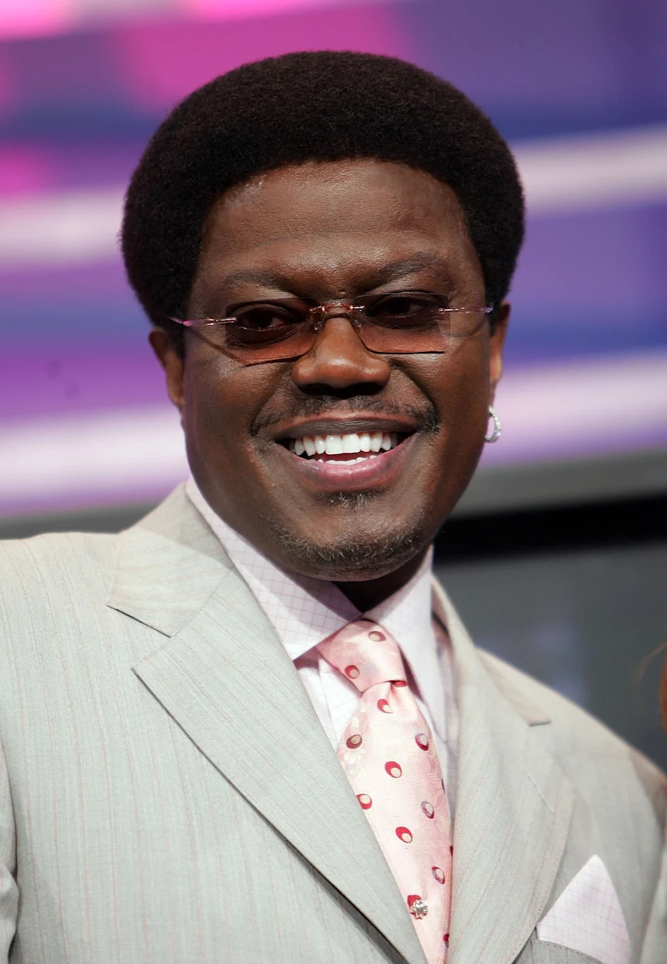 Bernie Mac in a suit and patterned tie with an afro and sunglasses smiles on a red carpet