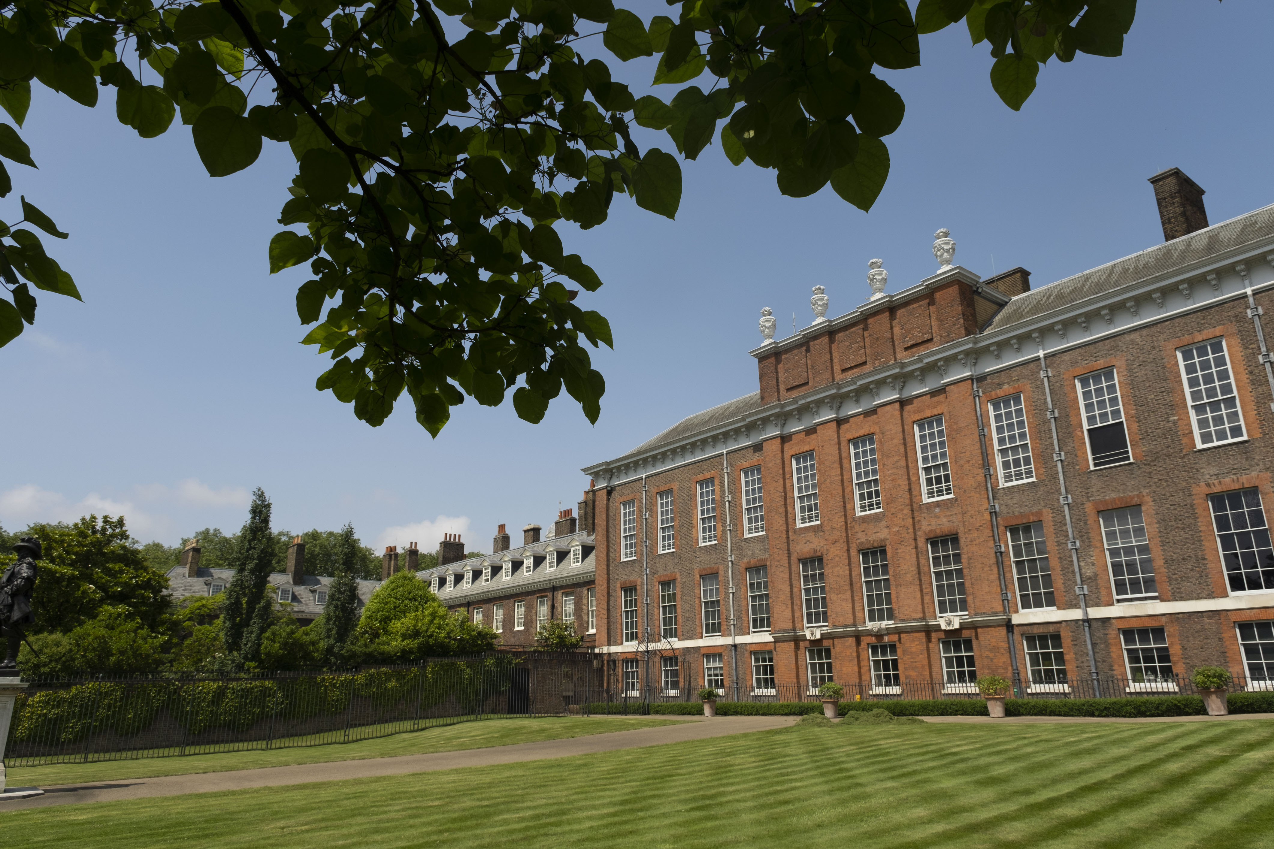 The brick exterior of Kensington Palace with a green lawn and a tree above it