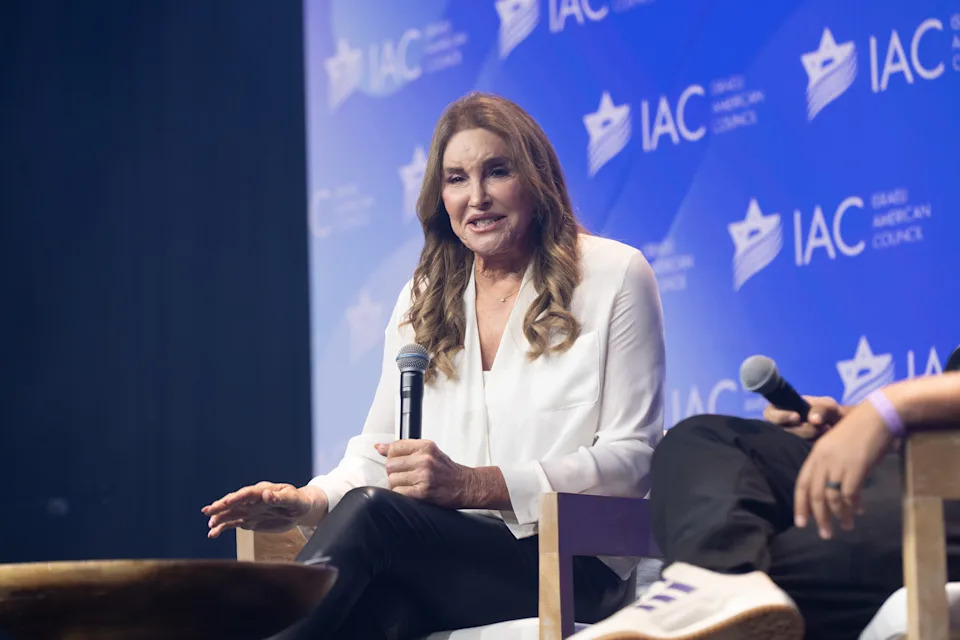 Caitlyn Jenner speaking on stage at a panel event, wearing a blouse and pants, holding a microphone with IAC branding in the background