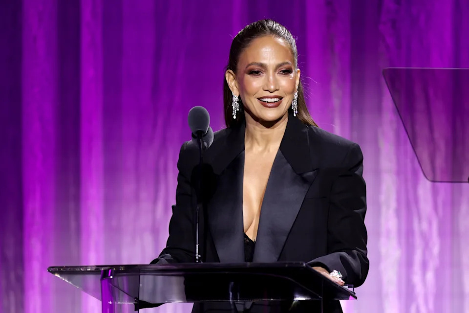 A person stands at a podium with a microphone, wearing a tailored black jacket and statement earrings, on a stage with a purple backdrop