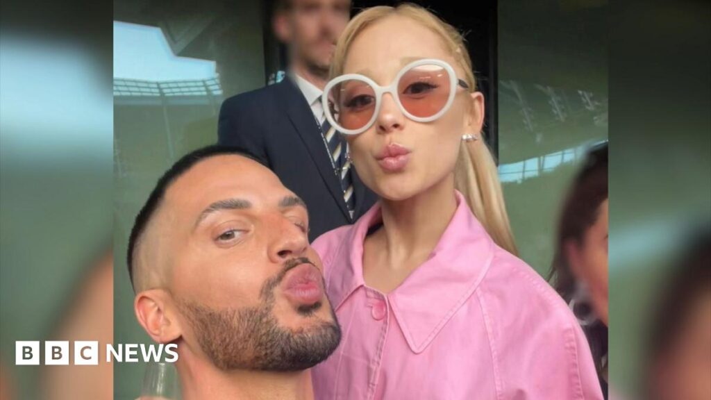 Carl Bembridge poses with the singer Ariana Grande. Carl and Ariana are both pouting and looking at the camera for a selfie. Ariana wears a pink jacket and white rimmed sunglasses.