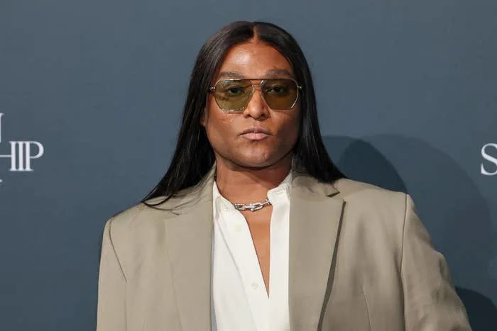 A person in a stylish beige suit with sunglasses poses confidently at a celebrity event