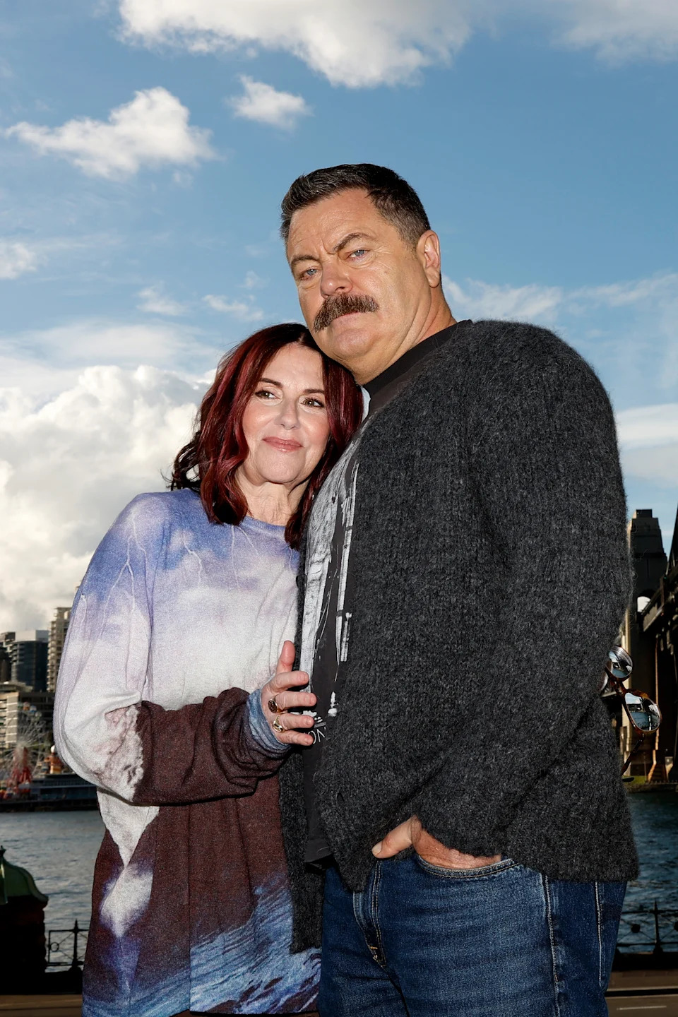 Two people standing closely outdoors, with one wearing a graphic tee and cardigan, and the other in a patterned sweater. Urban skyline in the background