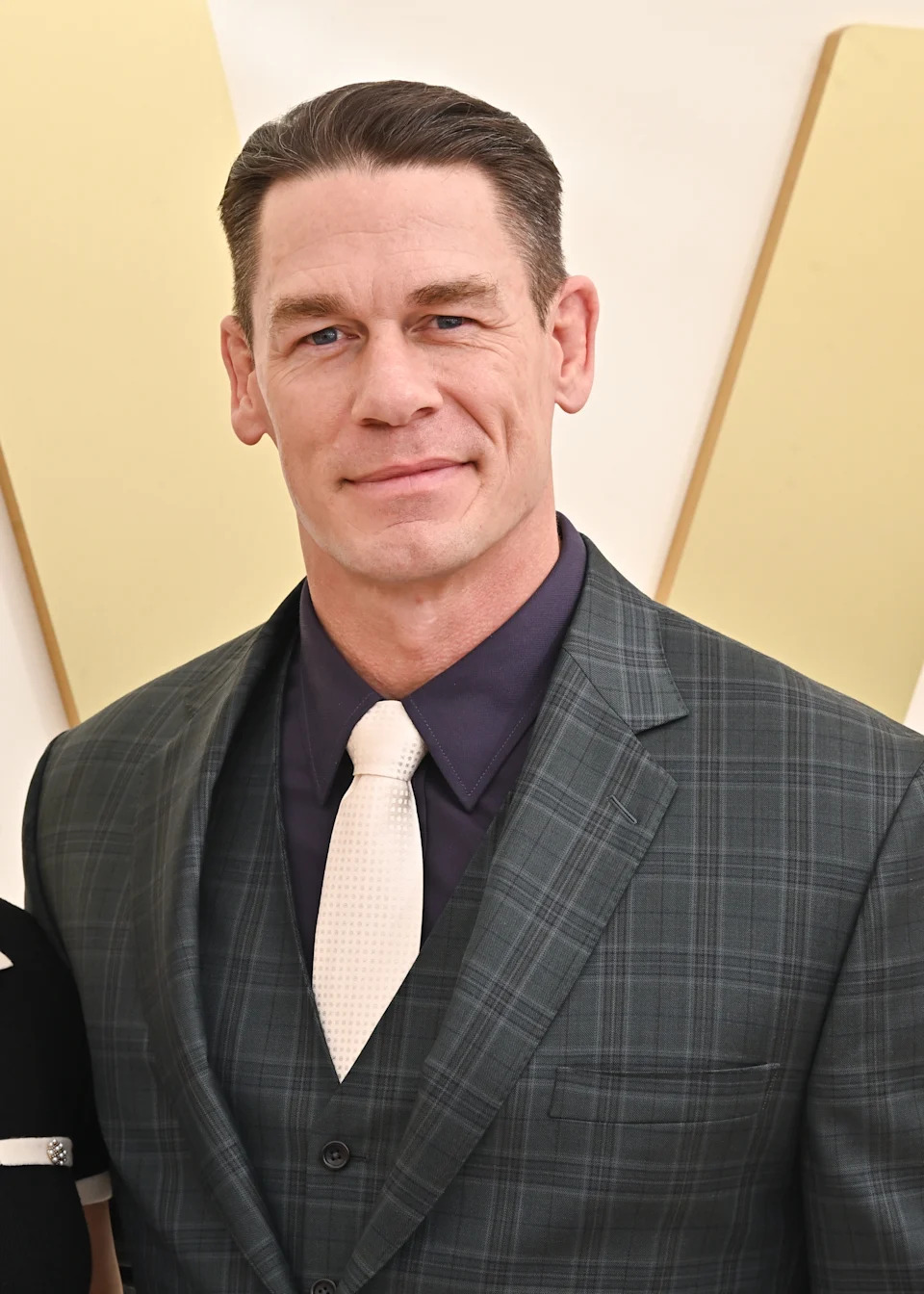 Person in checkered suit and white tie on a red carpet, smiling at the camera