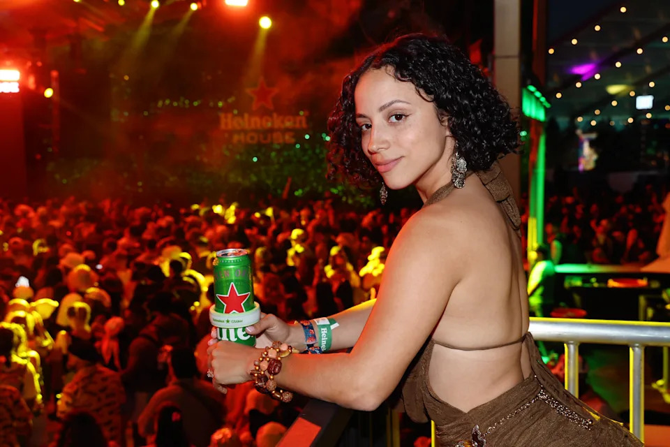 Kiana Madeira at a crowded event holds a drink, wearing a stylish halter top dress with curly hair