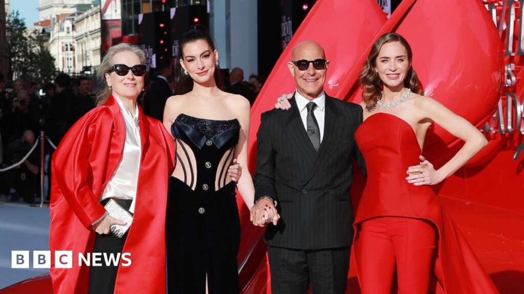 Meryl Streep, Anne Hathaway, Stanley Tucci and Emily Blunt pose on the red carpet in London. Streep is wearing sunglasses, a long, red, satin jacket, a cream blouse and black trousers. Hathaway has her hair up and is wearing a black embellished corset. Tucci is wearing a pinstriped black suit, white shirt and tie, along with sunglasses. Blunt has a red two-piece on an a jewelled necklace.