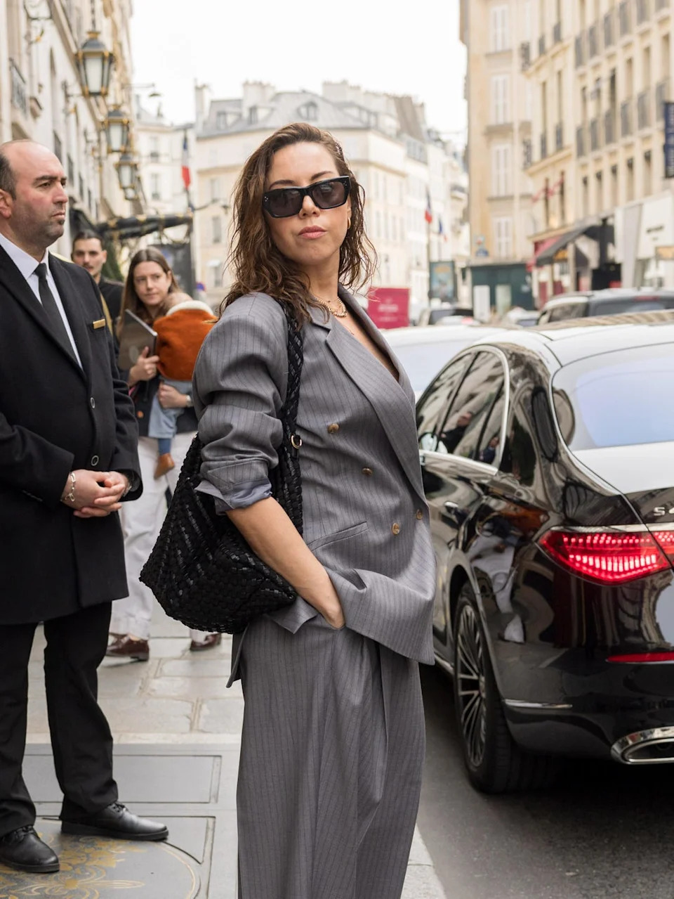 person in a stylish gray outfit on a city street near a luxury car
