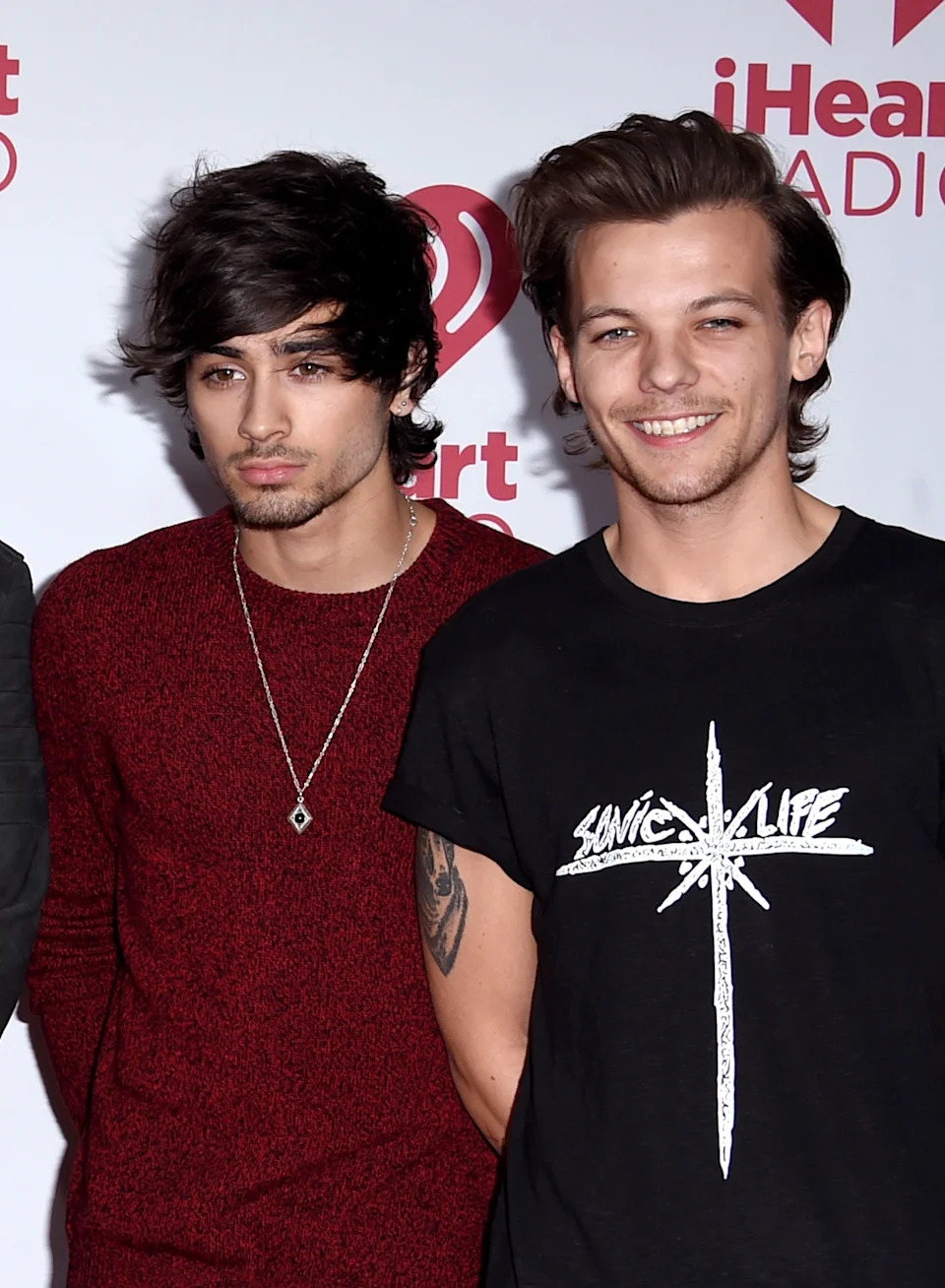 Zayn Malik wearing a sweater and Louis Tomlinson in a graphic t-shirt, smiling in front of a media backdrop