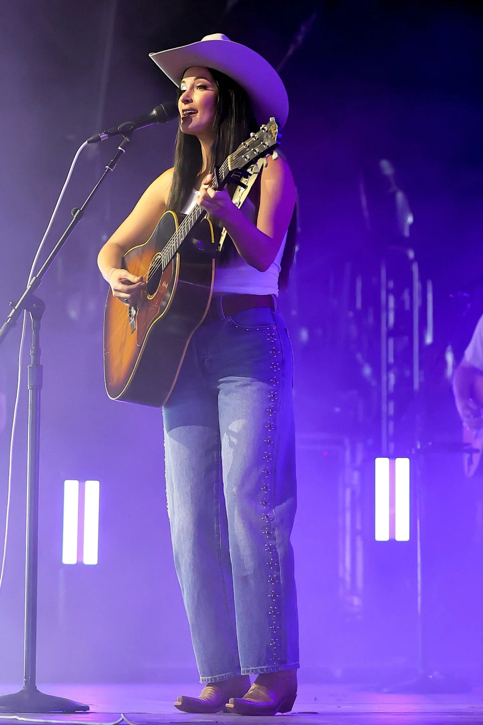 Kacey Musgraves on stage, wearing a cowboy hat, sleeveless top, and embroidered jeans, playing guitar and singing into a microphone