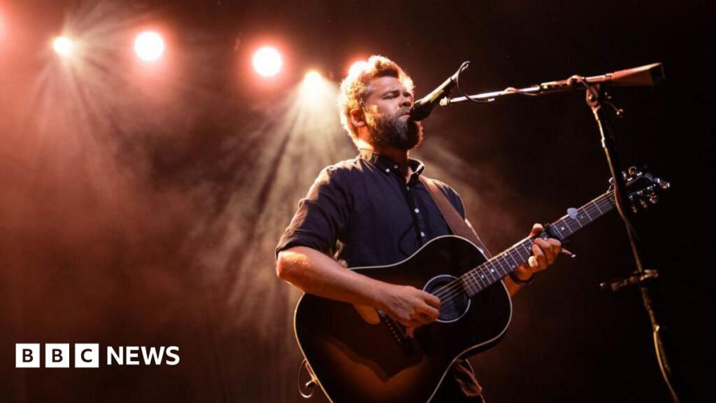 A man plays the guitar on stage. Behind him are red lights.