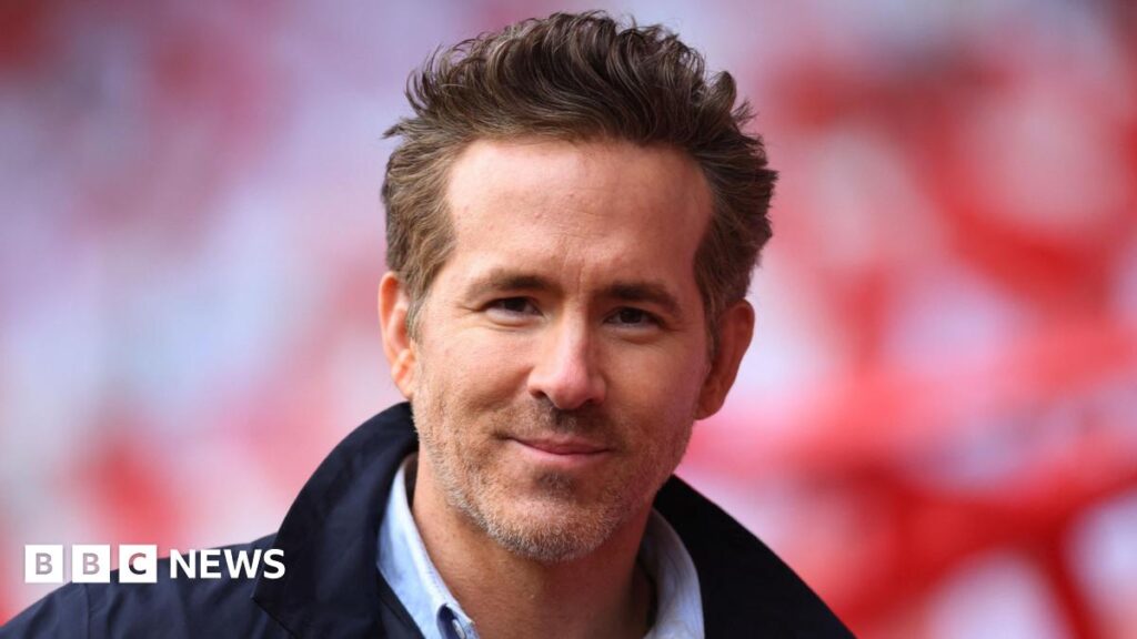 Ryan Reynolds looking directly at the camera, smiling. He has short light brown hair and facial hair. He is wearing a dark navy jacket and a shirt.