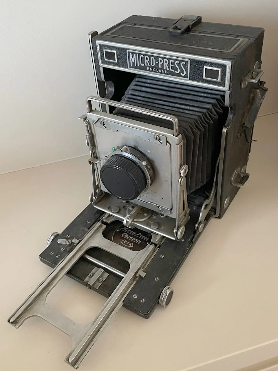 A Micro-Press England large format camera, its bellows fully extended and film holder rails deployed, photographed against a plain pale background.