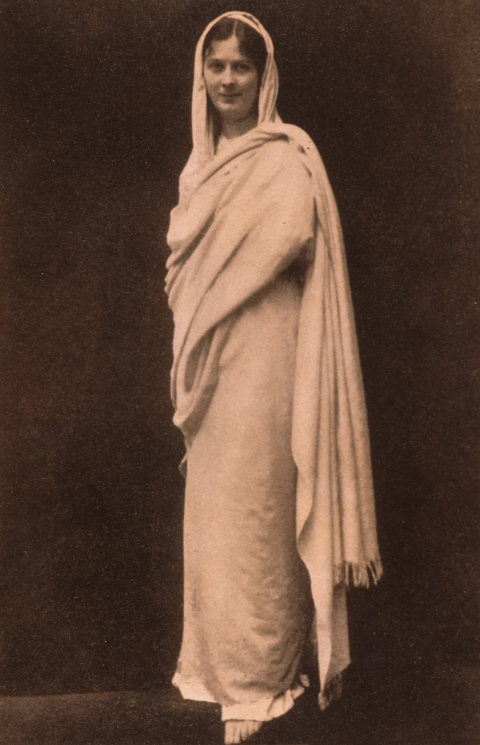 Isadora Duncan in traditional draped attire, standing with a serene expression against a plain backdrop