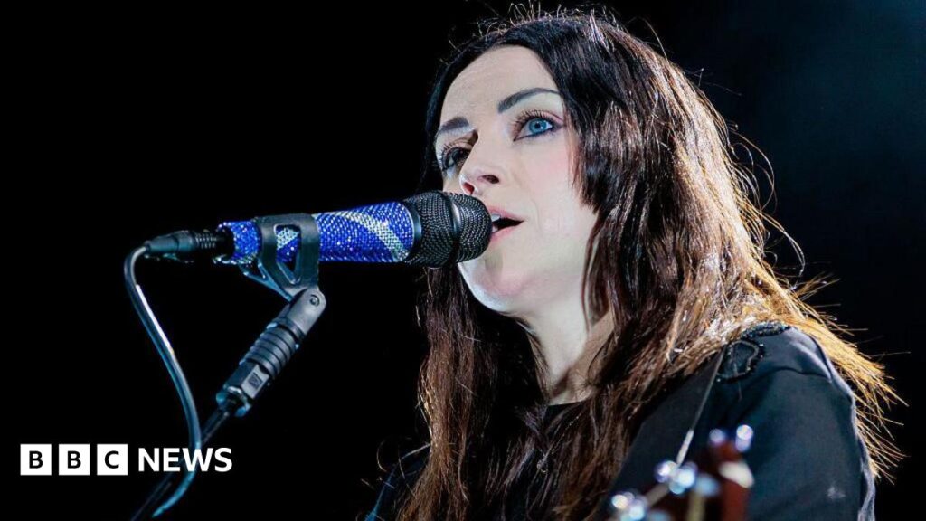 Amy Macdonald performs onstage, singing into a microphone. She has long dark hair and is playing a guitar