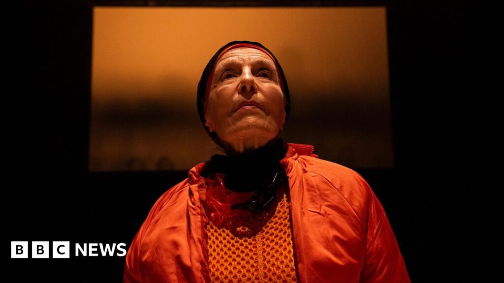 A woman wearing orange clothing and black hood looking out in the distance. The background is dark with a large orange picture.