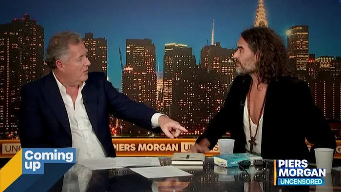 Piers Morgan and Russell Brand, seated at a news desk, engaged in a lively discussion on "Piers Morgan Uncensored" with a city skyline backdrop