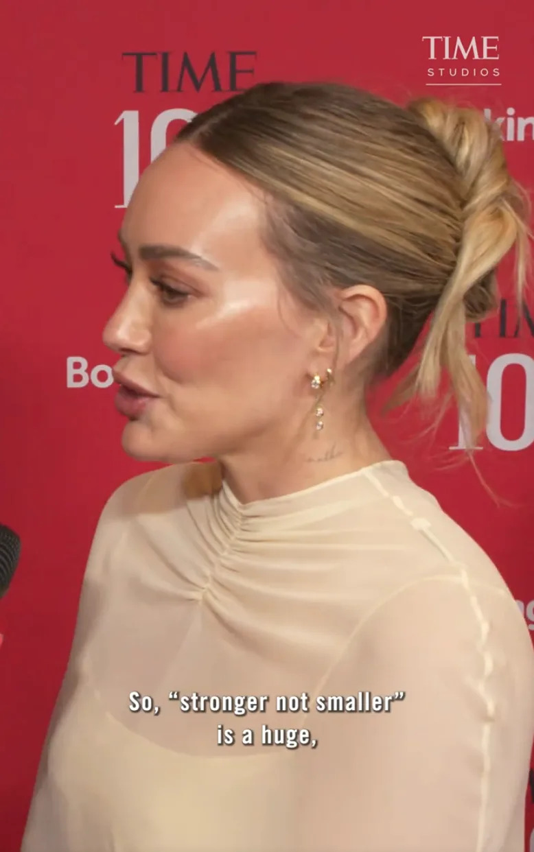 Person with hair up, wearing a sheer, elegant top, speaking at a red carpet event. Text on image: "So, 'stronger not smaller' is a huge..."