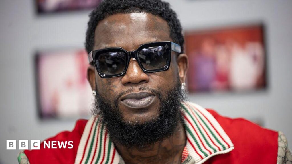 A close-up of Gucci Mane. He wears sunglasses and a red shirt.