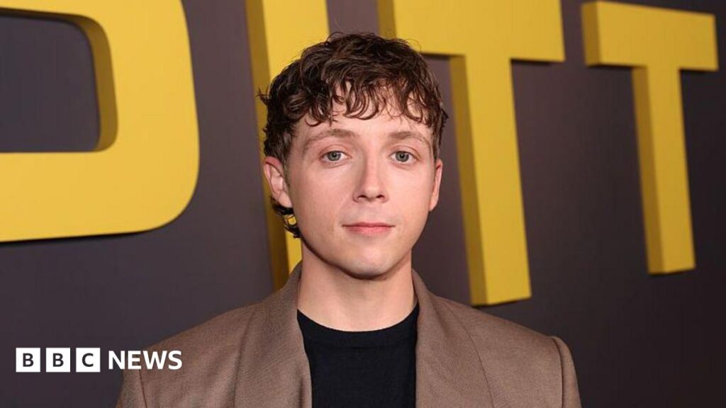 Nathan sitting in a pub with a drink. He is wearing a blue tracksuit top and has his arms crossed. He has a short beard and dark brown hair.