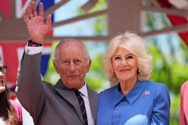 The King waves as he stands next to the Queen, who is wearing blue