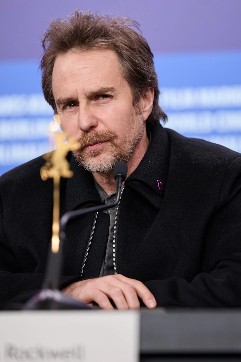 Person at a press conference wearing a dark jacket and shirt, sitting behind a microphone, with an award visible in the foreground