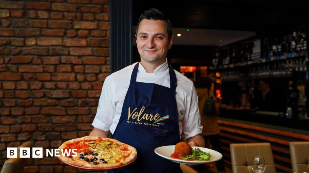 Jason Maimone smiles as he holds a pizza and another Italian dish in his restuarant. He wears a blue apron with the Italian tricolour over his chef whites.