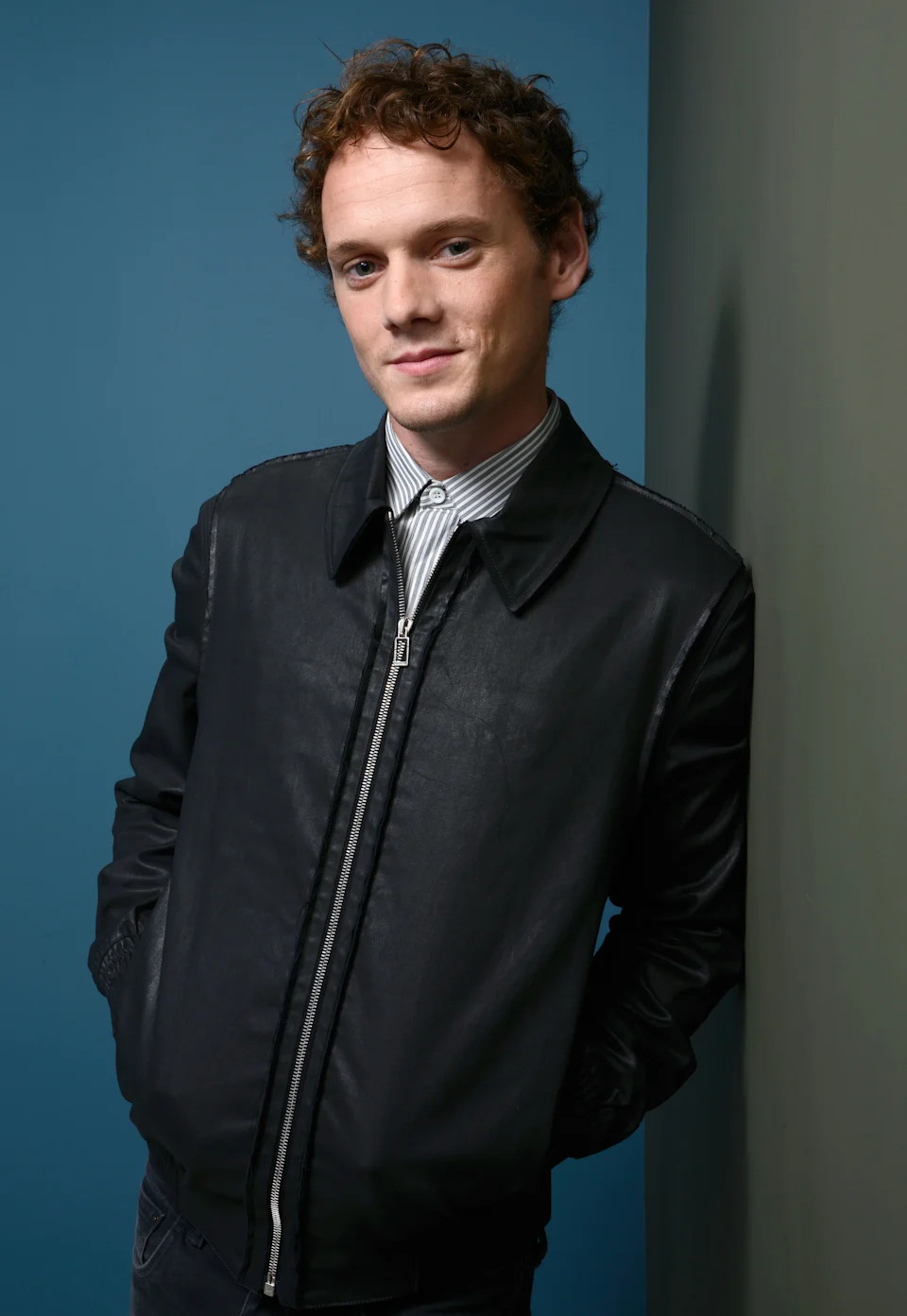 Anton Yelchin in a casual bomber jacket poses against a wall, hands in pockets, with a relaxed expression