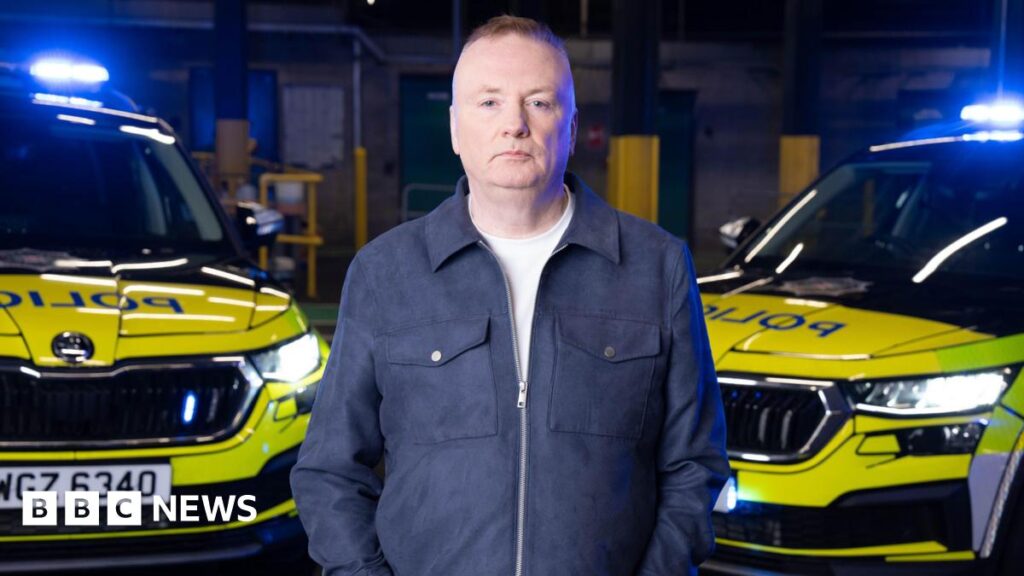 Stephen Nolan looking at the camera, he has brown hair. He is wearing a navy jacket that is zipped up halfway with a white t-shirt under it. Behind him are two police cars that have blue lights on.