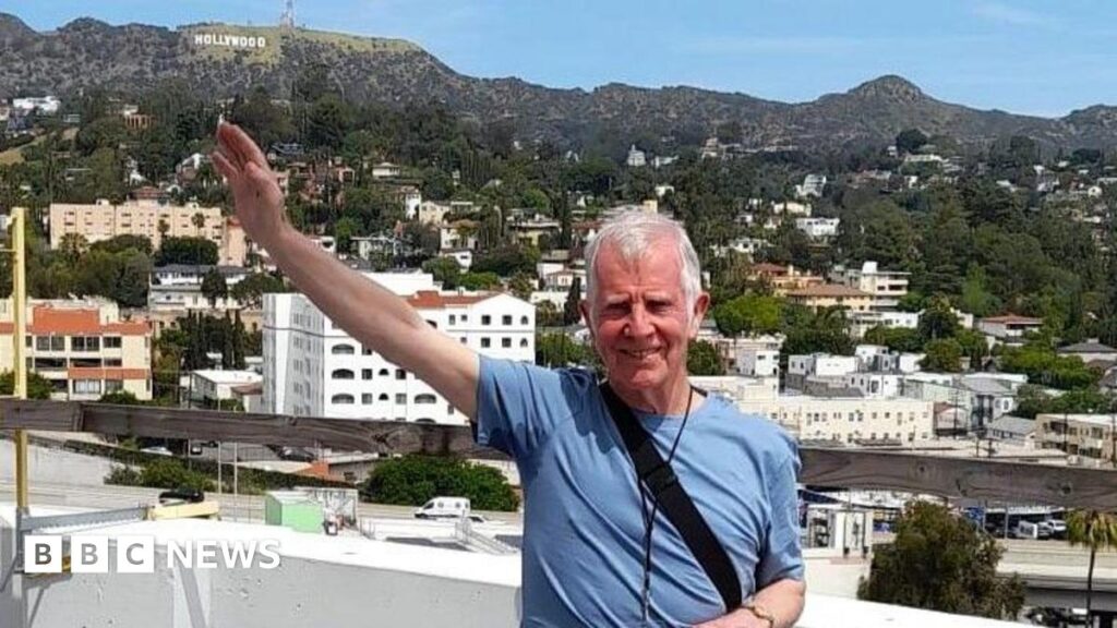 Jim, a mature man with white short hair and wearing a light blue T-shirt raises his arm towards the "Hollywood" sign as he checks out the roof on his tour of the Capitol Records building.