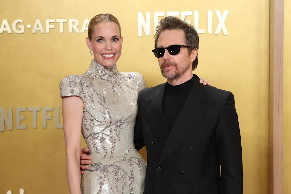 Two celebrities on a red carpet: one in an elegant silver gown with puff sleeves, the other in a suit and sunglasses